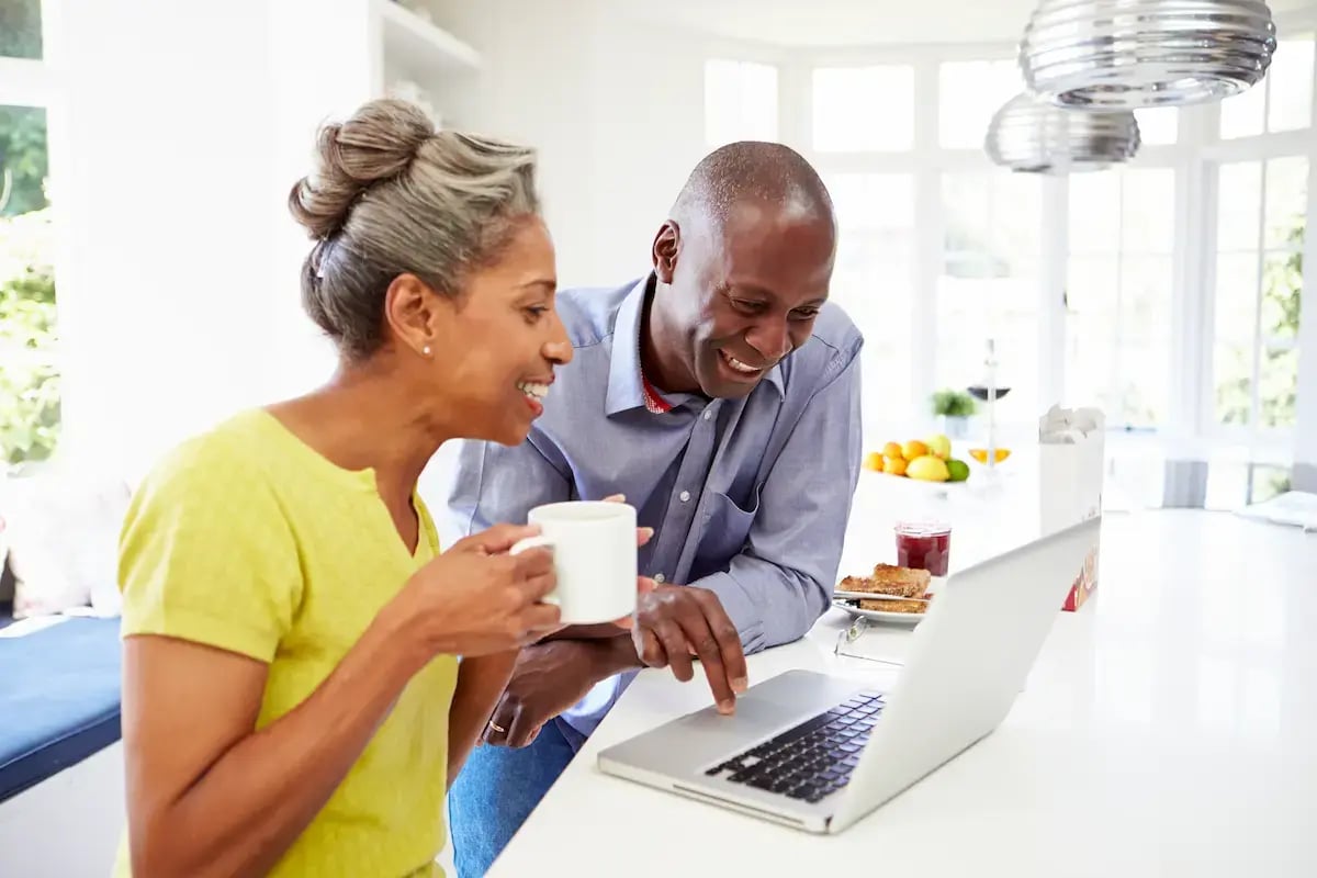 The image shows two individuals in an indoor setting, with one person using a laptop and the other holding a mug.